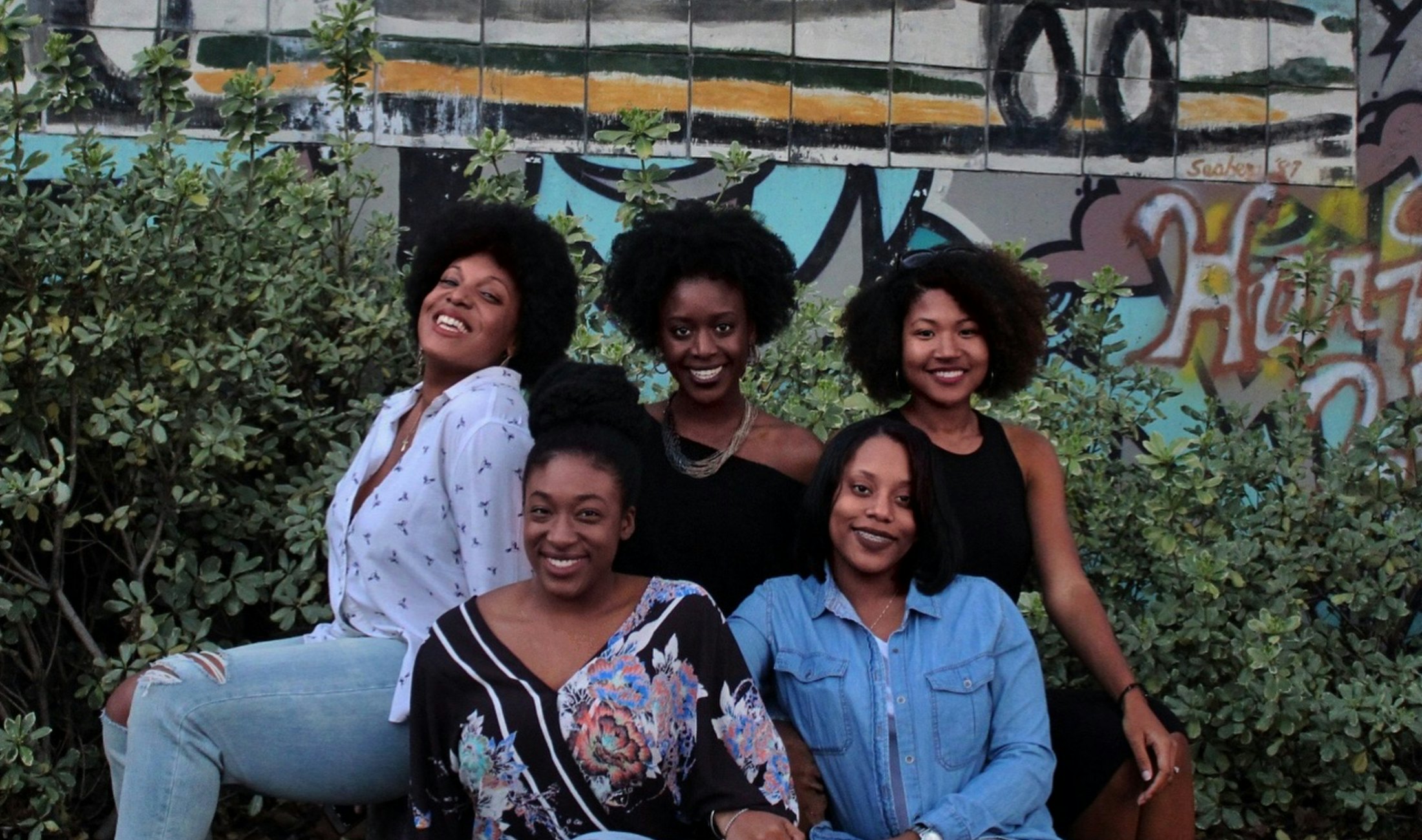 Five Black women sitting together in front of a mural, smiling